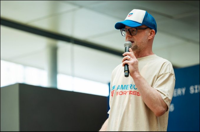 A man wearing a baseball cap, glasses, and a 'AMERICA FOR FEST' t-shirt speaks into a microphone at what appears to be a rally or event.