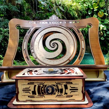 A wooden art piece or decorative box featuring a spiral design, displayed outdoors with greenery in the background.