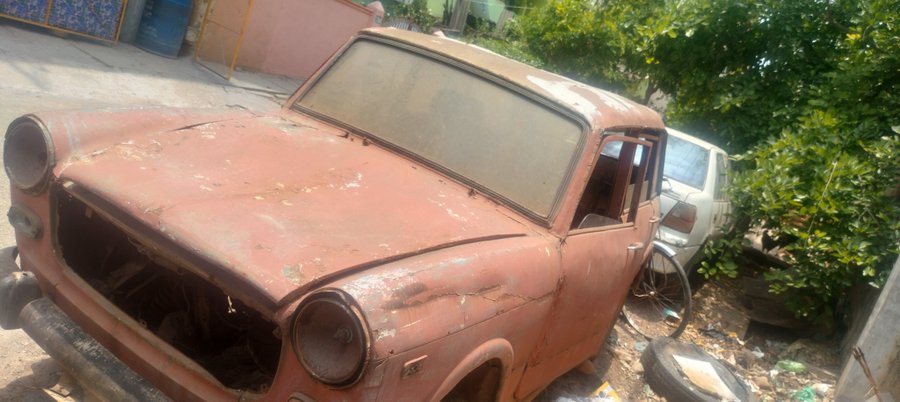 An abandoned, rusty red car with peeling paint parked in a cluttered area with vegetation and tires nearby.