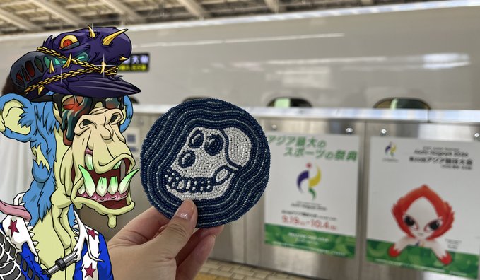 A person holds up a crocheted patch featuring a skull design, juxtaposed against a cartoon zombie character, with Japanese Olympic and Paralympic posters visible in the background.