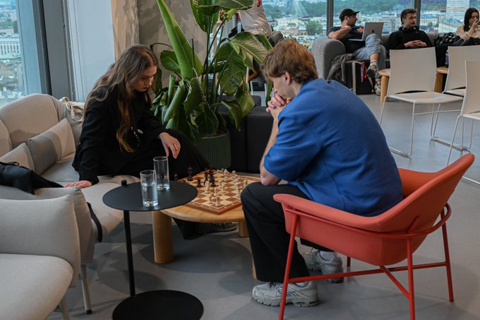 Two people sit across from each other playing chess at a wooden table in a lounge area with a large potted plant, with two glasses of water on a side table.