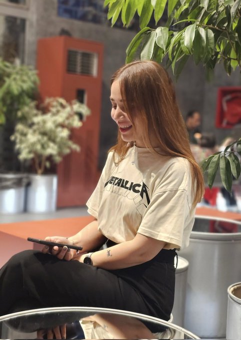 A young woman with long auburn hair smiles while looking at her phone, wearing a cream-colored Metallica t-shirt and a small wrist tattoo. The image complements the post by showcasing the artist, who is announcing a price drop on her digital artwork "Connected" (1/1, 0.03 ETH) available on Transient Labs.