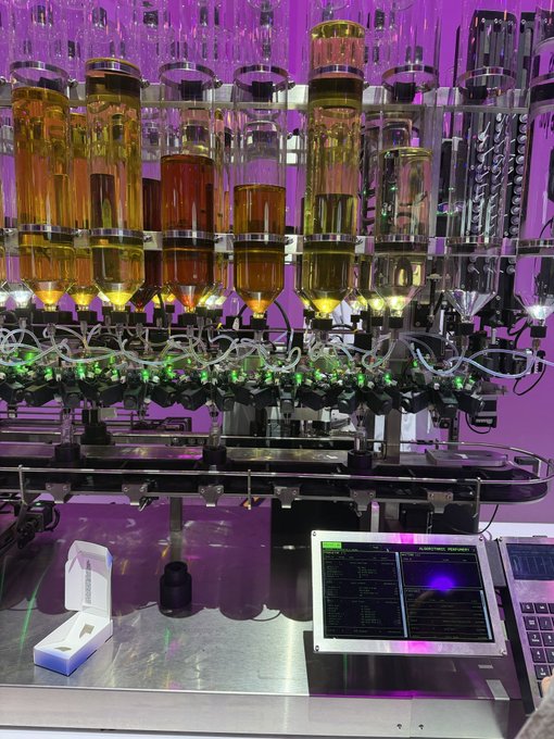 Industrial liquid filling machinery with multiple transparent bottles containing golden-amber liquid on a rotating production line, featuring illuminated nozzles and a digital touchscreen control panel on a stainless steel surface.