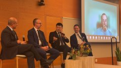 Four men in suits sit on stage during what appears to be a panel discussion, with a large screen behind them showing a video call participant's face.