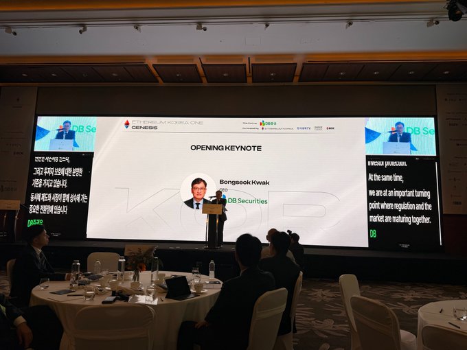 Bongseok Kwak of DB Securities delivers an opening keynote at "Ethereum Korea One: Genesis," with screens highlighting themes of regulation and market evolution in Korean and English. The event caption reflects gratitude from Optimism for DB Securities' partnership in bringing real-world assets onchain in Korea.
