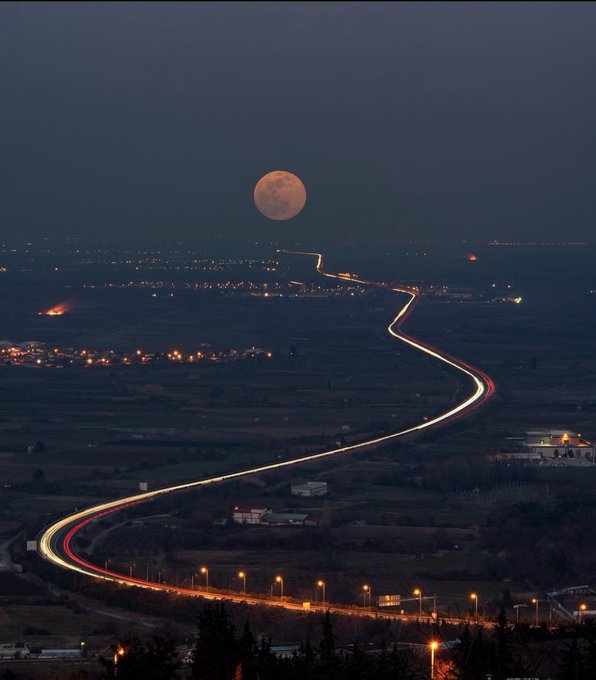 A nighttime long-exposure photograph showing a winding highway with light trails from vehicles, illuminated street lamps, and a full moon hanging in the dark sky.