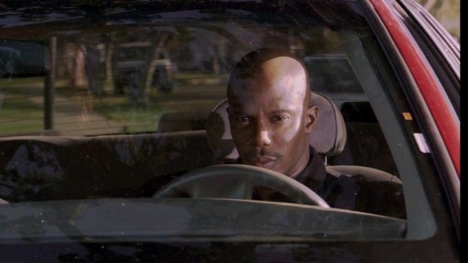A man with a serious expression sits behind the steering wheel of a car, viewed through the windshield on what appears to be a sunny day.