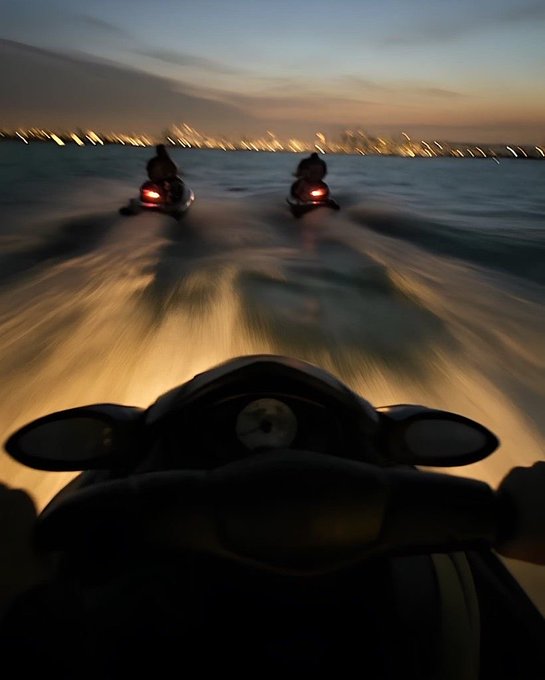 A dynamic first-person view from a jet ski at dusk, showing two other riders ahead on the water with a blurred city skyline in the distance.