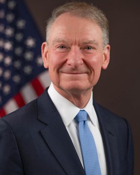 A formal portrait shows an older man with gray hair wearing a dark suit and light blue tie, smiling slightly with an American flag visible behind him. The image likely depicts Paul Atkins, the current SEC Chairman, whose agency proposed the new crypto custody rules allowing investment advisers to hold clients' digital assets themselves when qualified custodians are unavailable.