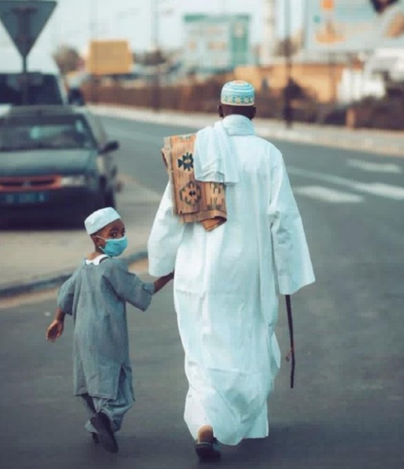 A touching photograph of a man in traditional white Islamic attire walking down a street, holding hands with a young boy wearing a face mask and traditional clothing.