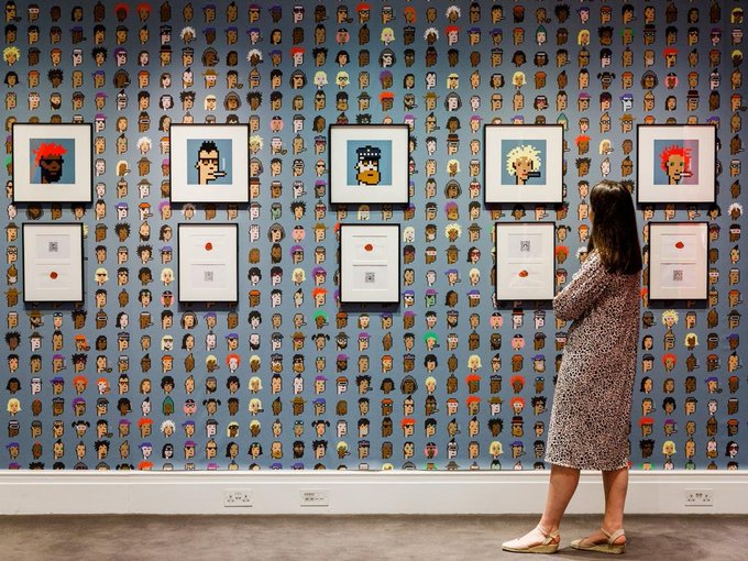 A woman in a leopard print dress viewing a gallery exhibition featuring framed CryptoPunks NFT artwork mounted on wallpaper covered with a grid pattern of small pixel-art character icons.