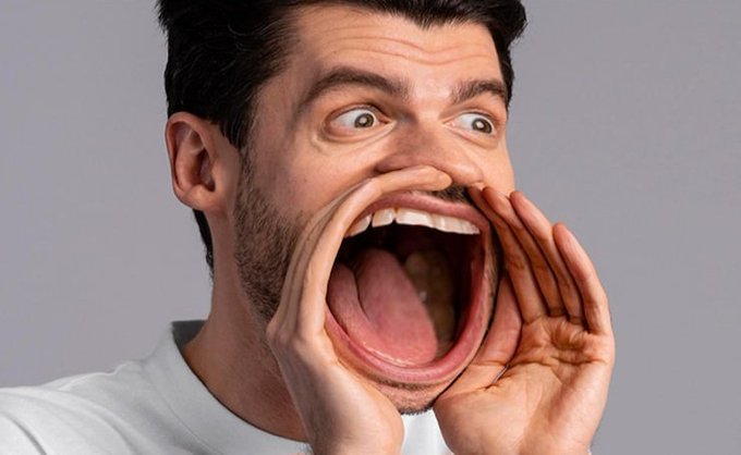 A man with dark hair and a beard screaming with his mouth wide open, hands cupped around his face against a gray background.