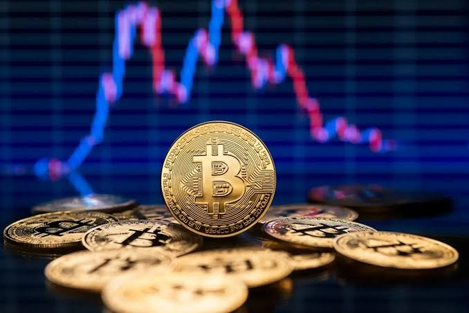 Several gold-colored Bitcoin tokens are stacked in the foreground against a backdrop of a red and blue stock market price chart.