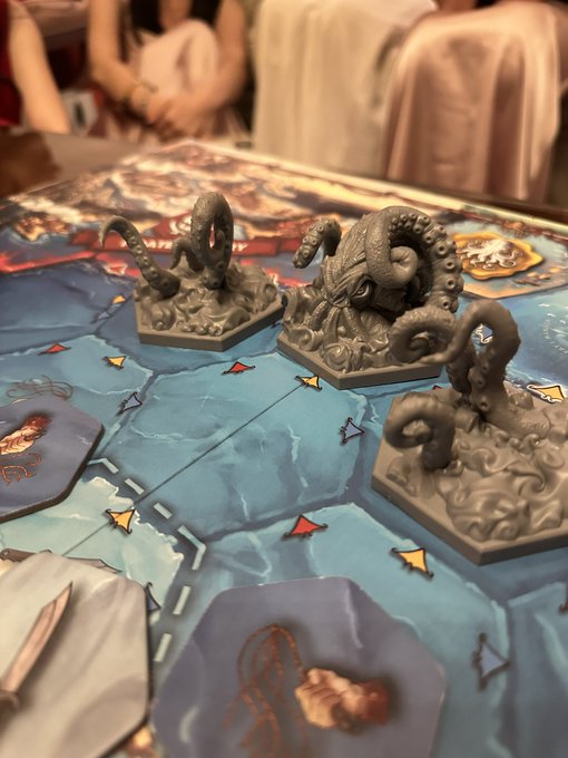 A close-up of a board game in progress on a table, featuring three grey tentacle creature miniatures placed on a blue ocean-themed game board, with people sitting in the blurred background.