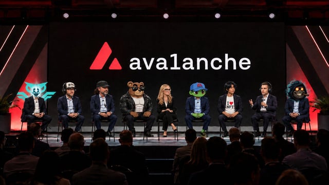 The image features a group of people sitting around a stage, with a large screen behind them displaying the words "AVALANCHE". There are at least eight people visible in the scene, all dressed in suits and ties. The audience is seated on chairs arranged in two rows, facing the stage where they appear to be engaged in a discussion or presentation related to the company name "AVALANCHE".