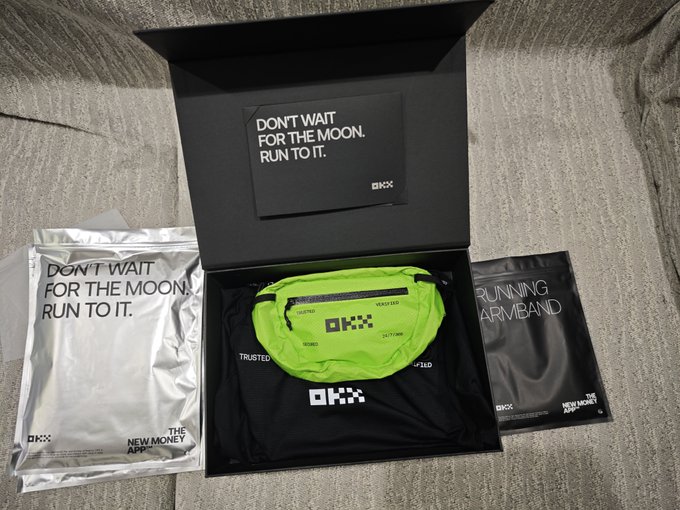 An unboxed OKX merchandise package sits on a bed, containing a neon green running waist pack, a black t-shirt, silver packaging that reads 'DON'T WAIT FOR THE MOON. RUN TO IT.' and 'THE NEW MONEY APP', a matching note card, and a running armband.