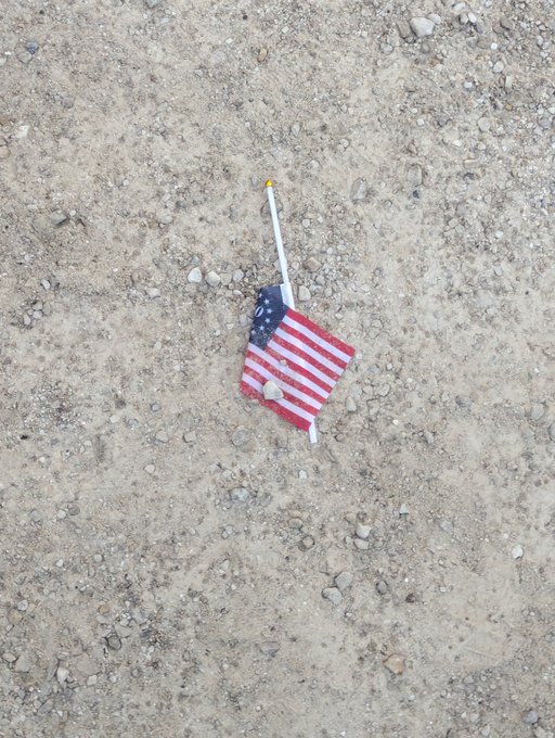 A small American flag lying on the ground on a patch of dirt and gravel.