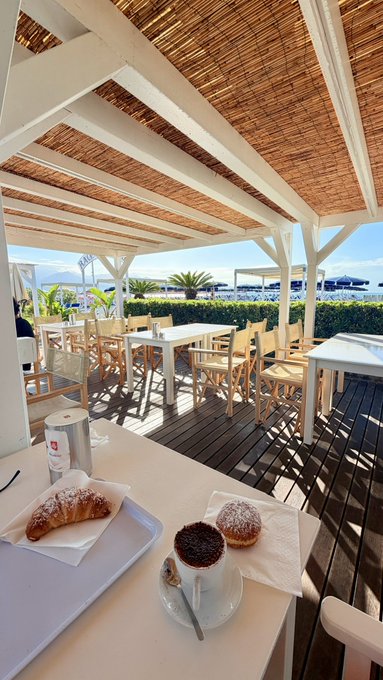 A tray with a chocolate croissant, two small cakes, and a coffee cup sits on a white table under a wooden pergola overlooking an outdoor patio and beach.