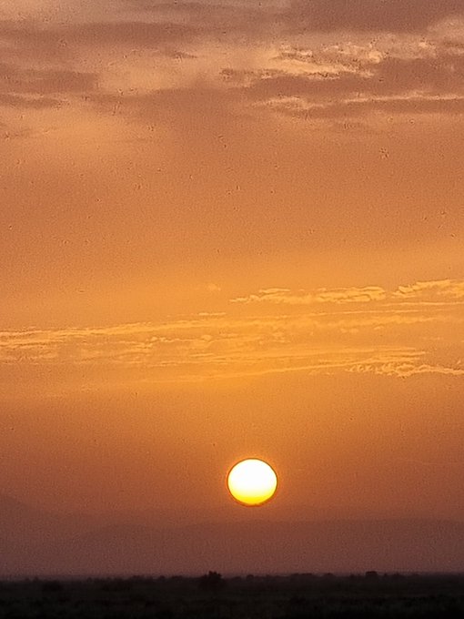 The image features a beautiful sunset with an orange sky, which serves as the backdrop for a large sun setting over a field of grass. The sun appears to be located near the horizon line, creating a stunning visual effect. In the background, there is a mountain range visible in the distance, adding depth and scale to the scene.