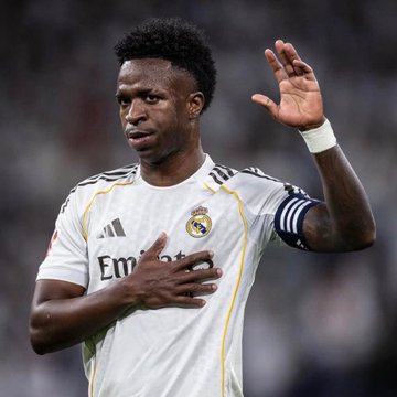 A photograph of soccer player Vinícius Júnior in a white Real Madrid jersey, making a hand gesture while wearing the captain's armband.