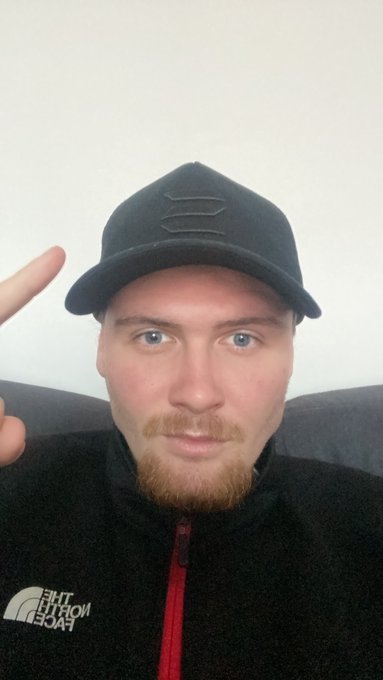 A selfie of a man with blue eyes and a blonde goatee wearing a black The North Face jacket and a black cap with an embossed logo, holding up his hand in a gesture.