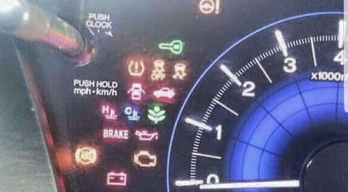 Close-up of a car's instrument panel showing the odometer/tachometer (reading near 0 x1000 RPM) with various illuminated warning lights including tire pressure, brake, ABS, battery, and check engine indicators.