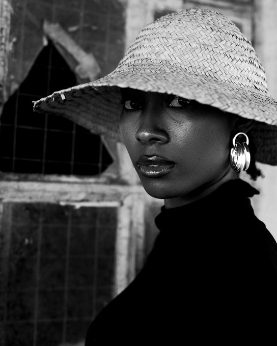 A black-and-white close-up portrait of a woman wearing a wide-brimmed straw hat, large hoop earrings, and a dark top, looking over her shoulder at the camera.
