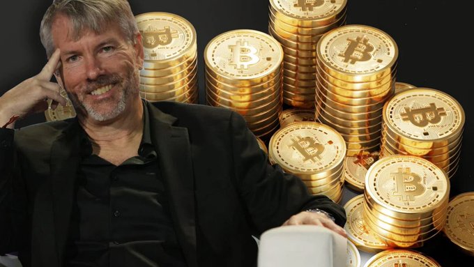 A man with a grey beard in a black shirt and jacket sits smiling with his hand on his head, posed in front of a large background of stacked gold Bitcoin tokens.