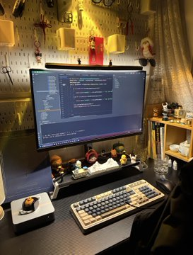 A desk setup featuring a large monitor displaying code, a mechanical keyboard, a mouse, and a pegboard wall decorated with small plush toys and hanging items.