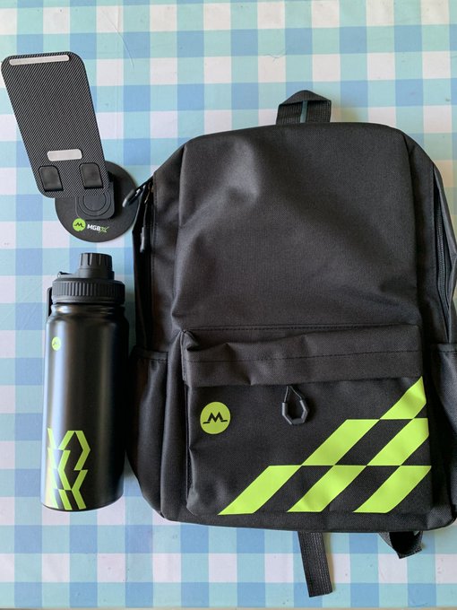 A black backpack with neon green "M" (MGBX) branding and matching water bottle are displayed on a blue gingham surface, representing the MGBX exchange merchandise received from the project team's promotional activity mentioned in the post about heading "to the moon" (去往月球).