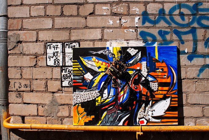 A vibrant street art painting in bold primary colors leaning against a graffiti-covered brick wall.