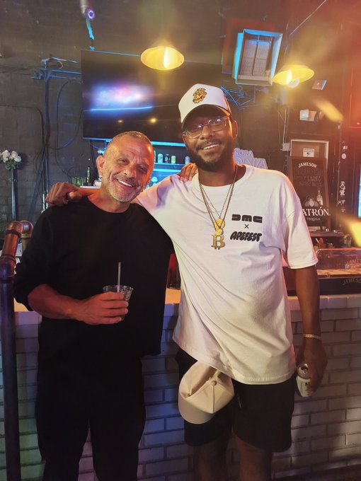 Two men posing together at a bar or lounge; the man on the left wears black and holds a drink, while the man on the right wears a white t-shirt with 'NBA x AMBASSY' text, a white cap, and gold chains.