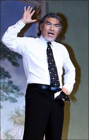 A man in a white dress shirt and black patterned tie raises his hand while standing in front of a painted backdrop.