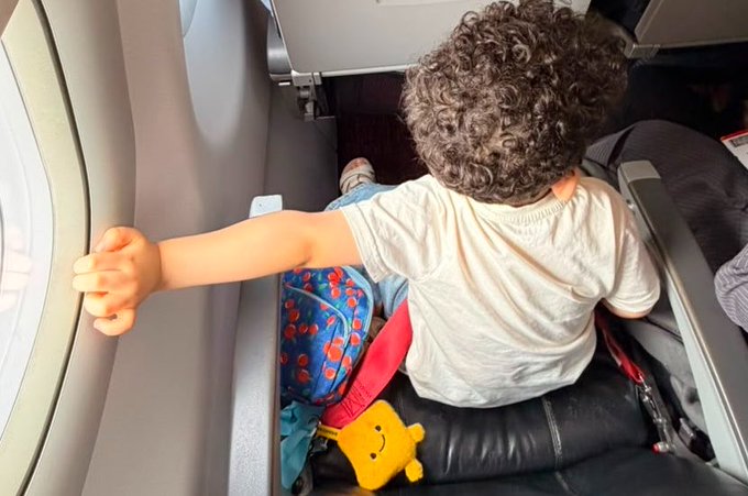 A curly-haired toddler sits on an airplane next to the window, wearing a white t-shirt and reaching out to touch the window frame, with a blue backpack featuring fish designs beside him and a yellow BiBi doll tucked at his side. The image illustrates the post's message about the child never going anywhere without his beloved BiBi companion, even bringing her on flights.