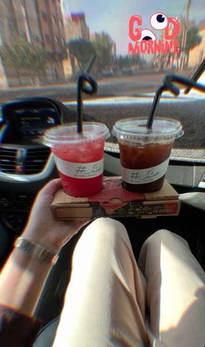 A first-person photo from inside a car showing two iced drinks (one pink, one iced coffee) on a book, with a 'GOD MORNING' graphic featuring cartoon eyes in the corner.