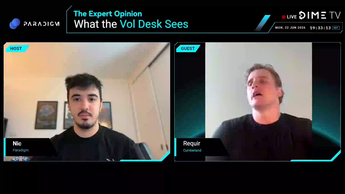 The image shows two men sitting behind computer screens, likely participating in an online video call or webinar. The screen displays the text "What the Vol. Desk Sees" along with a picture of a desk. One man appears to be speaking while the other is listening attentively. The background features a white wall and a poster on it.