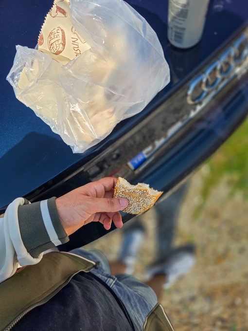 A hand holds a half-eaten sesame bagel near the open trunk of a blue car, with a Nissin cup noodle package and a white bottle resting on the trunk lid. The modest meal of cheap, on-the-go food paired with the "crypto victim" caption humorously reflects the bare-bones lifestyle often joked about among those who have lost money in the crypto market.