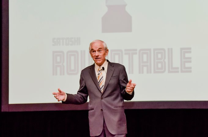 An older man in a suit and striped tie speaks on stage with open arms in front of a screen displaying 'SATOSHI ROUNDTABLE'.