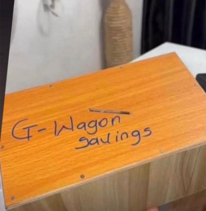 A wooden donation box labeled "G-Wagon Savings" in marker sits on a table, humorously representing the user's crypto investment goal—hoping their digital gains will eventually fund the purchase of a luxury Mercedes G-Wagon.