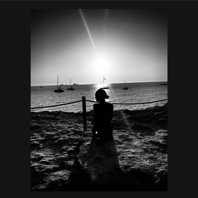 A black-and-white photograph captures the silhouette of a person wearing a cap sitting on a sandy beach at sunset, gazing out at sailboats on the water with the sun casting a bright reflection.