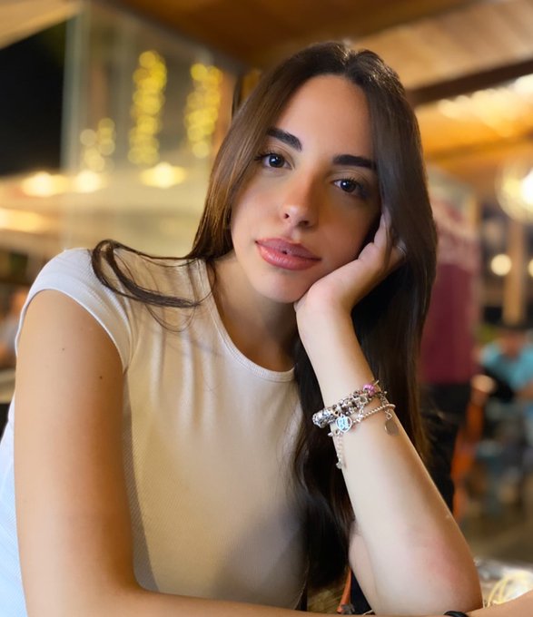 The image features a young woman sitting at a table, wearing a white shirt with a bracelet around her wrist. She has long brown hair that falls over her shoulders as she looks directly at the camera. The background of the photo shows other people present in the scene, but they are not the main focus of the image.
