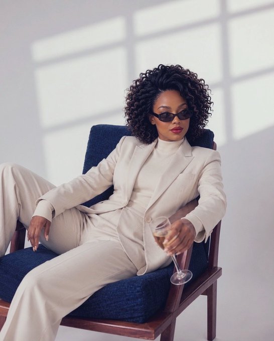 A stylish Black woman with voluminous curly hair wearing a cream-colored suit and small sunglasses, sitting confidently in a blue chair while holding a wine glass, with sunlight streaming through a large window behind her.