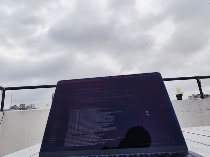 A laptop displaying a dark-themed code editor sits on an outdoor table against a cloudy sky, with the reflection of a person wearing a cap visible on the screen. The late-night coding session aligns with the post's mention of "token maxxin till 7am," suggesting a marathon of work on a token-related project before calling it a night.