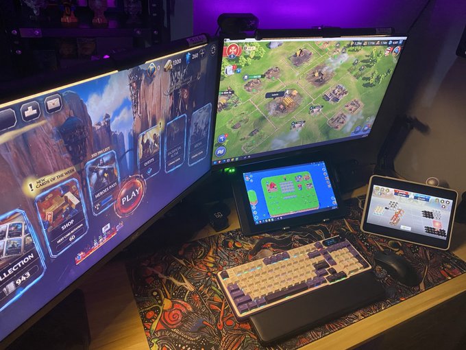 A gaming setup with two large monitors (one displaying a card game menu, the other a strategy game), a tablet and handheld device, a mechanical keyboard, and a mouse on a patterned desk mat.