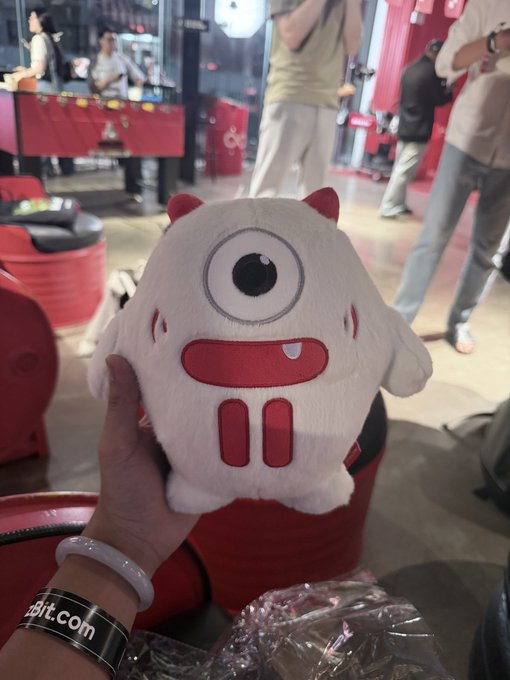A person holds a white plush toy with one eye and red horns at a red-themed event booth, while wearing a "bit.com" wristband, supporting the call to visit the @trondao booth for games and prizes.