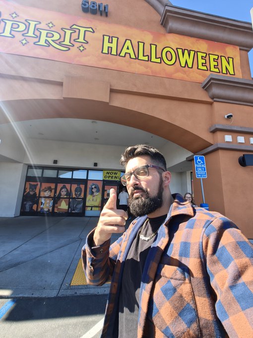 A bearded man wearing sunglasses and a plaid flannel shirt points at the camera while standing outside a Spirit Halloween store, with a 'NOW OPEN' sign visible in the background.