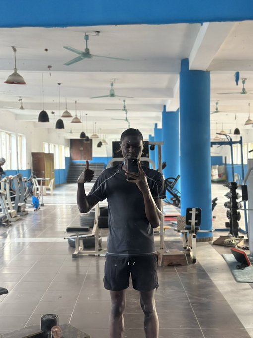 A man in dark athletic wear takes a mirror selfie in a gym, giving a thumbs-up, with exercise equipment, blue pillars, and ceiling fans visible in the background.