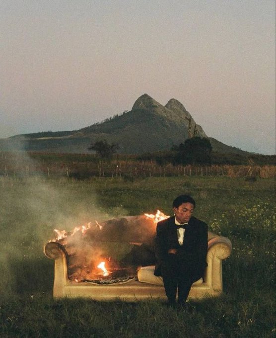 A cinematic photo of a Black man in a suit sitting on a burning couch in a field, with a large mountain in the background under a dusky sky.