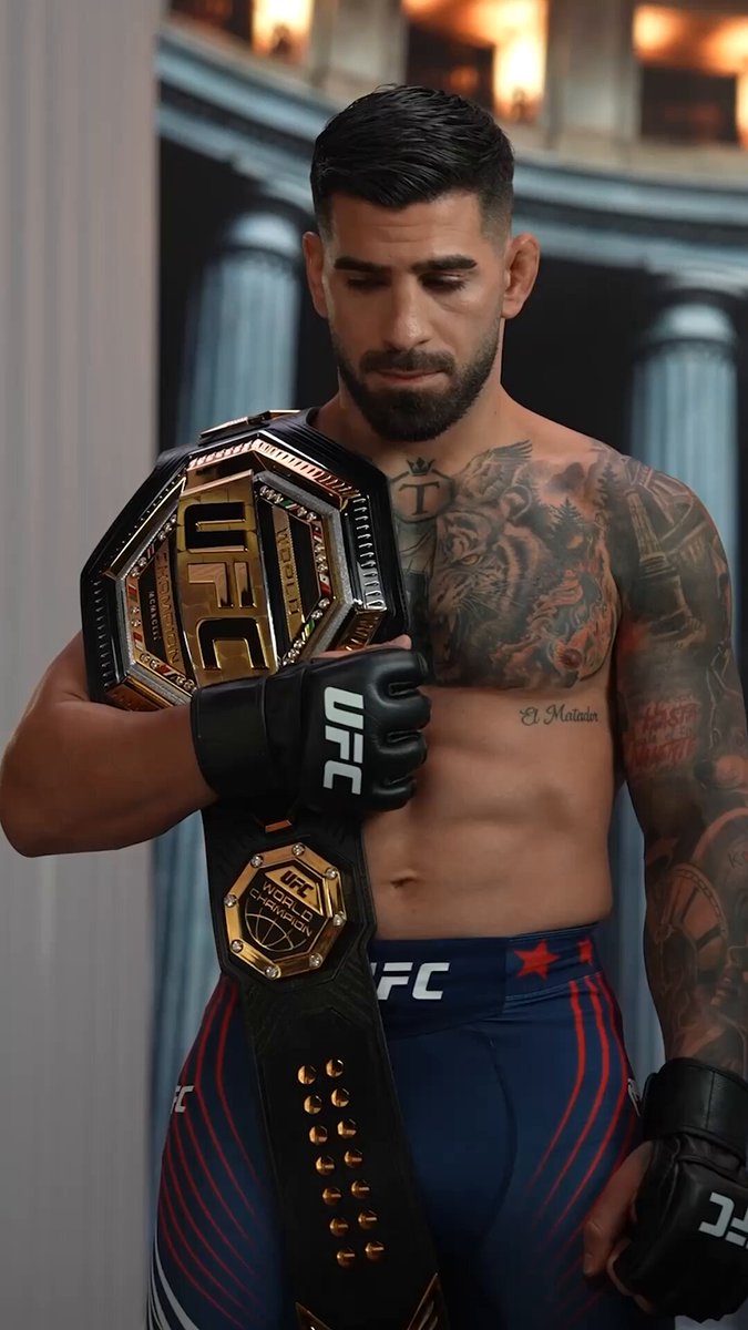 The image features a man holding a UFC championship belt, which has the UFC logo on it. The man appears to be posing for a photo or participating in an event related to the UFC. The background of the image shows a building with columns, suggesting that this could be a venue or a location associated with the UFC. The post also mentions that the event is scheduled for Saturday at 8 pm and will be live on social media platforms.
