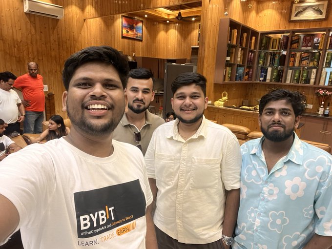 A group of four young men smile for a selfie at what appears to be a restaurant or lounge with wooden decor and framed pictures in the background, with one wearing a "BYBIT #TheCryptoHub and Gateway to Web3 LEARN. TRADE. EARN." t-shirt. The image captures the post's theme of colleagues meeting in person, likely during a crypto/Web3 event where they connected beyond their online interactions.